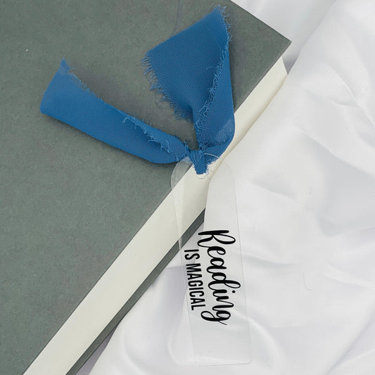 Liebevoll gestaltetes, abgerundetes Lesezeichen aus Acryl mit dunkelblauem Chiffonband und beklebt mit schwarzem Folienaufdruck "Reading is magical"
