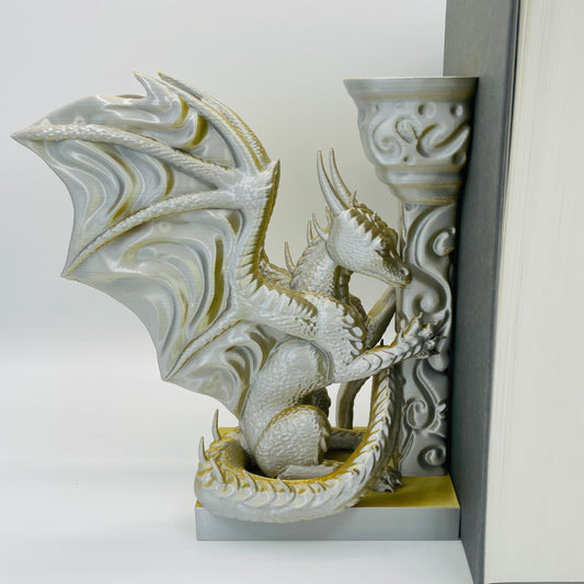 Detailliertere Ansicht einer 3D-gedruckten Buchstütze eines Drachens in Seitenansicht, angelehnt an eine Säule im Farbton Gold-Silber, die Buchstützte steht links vom Buch