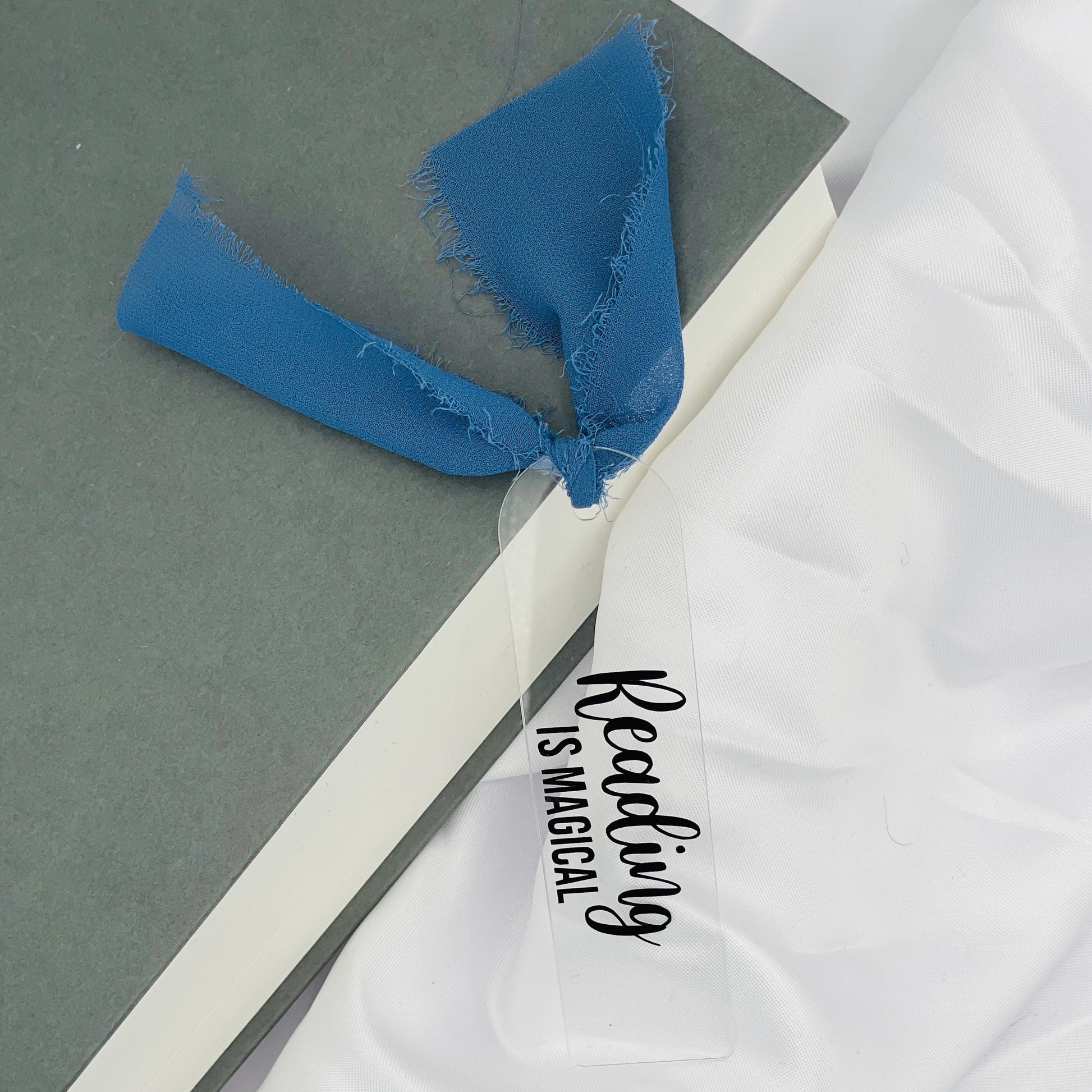 Liebevoll gestaltetes, abgerundetes Lesezeichen aus Acryl mit dunkelblauem Chiffonband und beklebt mit schwarzem Folienaufdruck "Reading is magical"