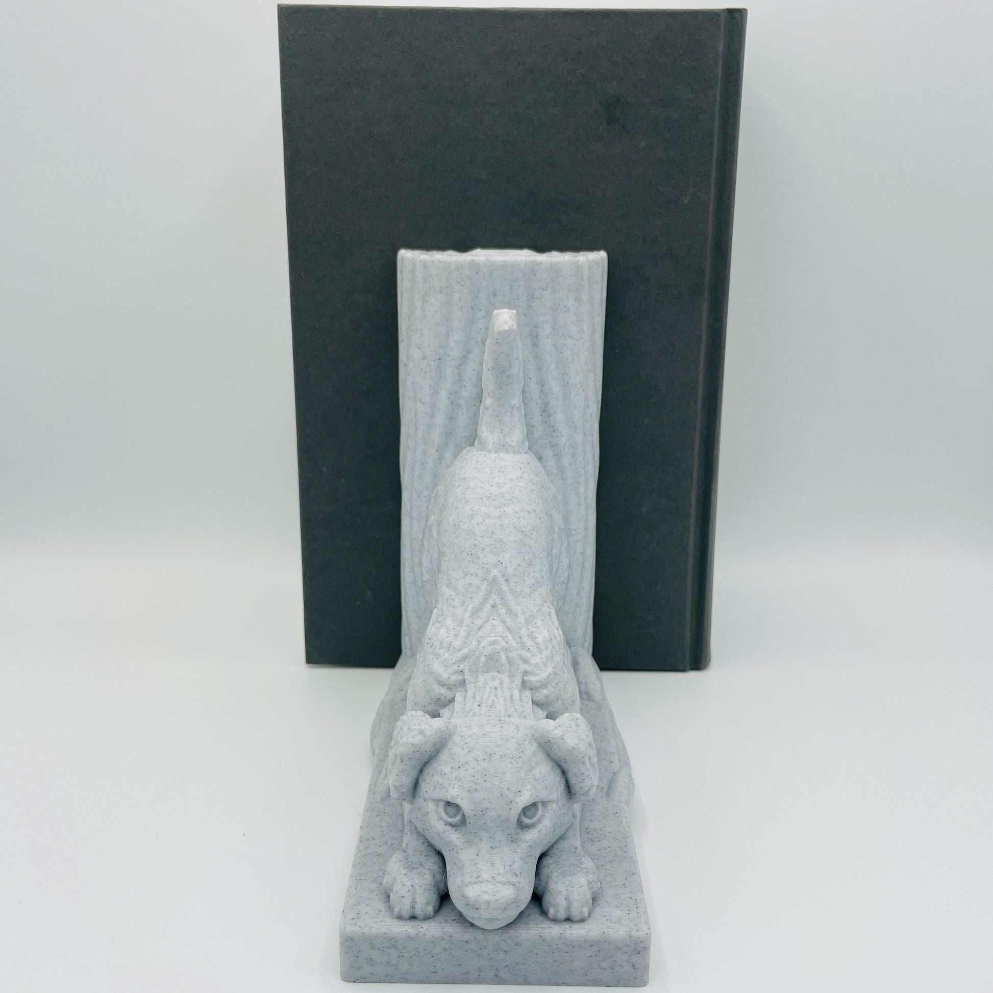 Frontalte Ansicht einer 3D-gedruckten Buchstütze eines süßen Hundes in Seitenansicht, der mit dem Oberkörper auf dem Boden lehnt im Farbton Marmor, die Buchstützte steht rechts vom Buch