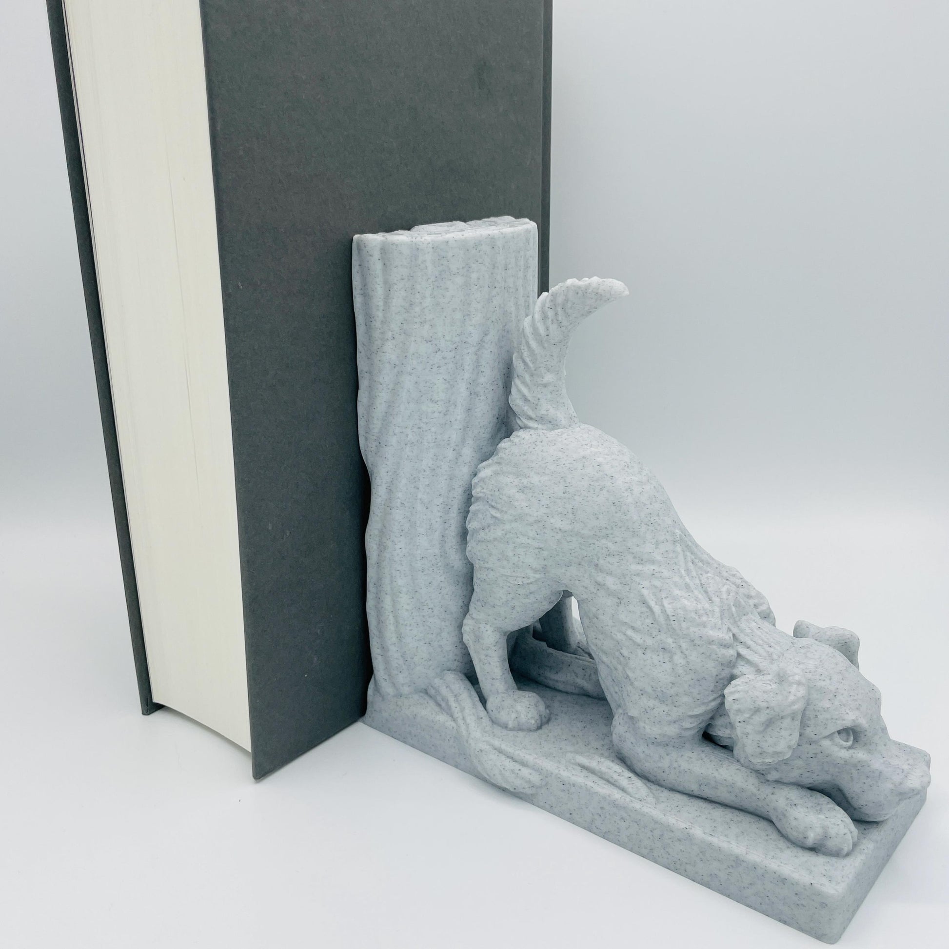 Diagonale Ansicht einer 3D-gedruckten Buchstütze eines süßen Hundes in Seitenansicht, der mit dem Oberkörper auf dem Boden lehnt im Farbton Marmor, die Buchstützte steht rechts vom Buch