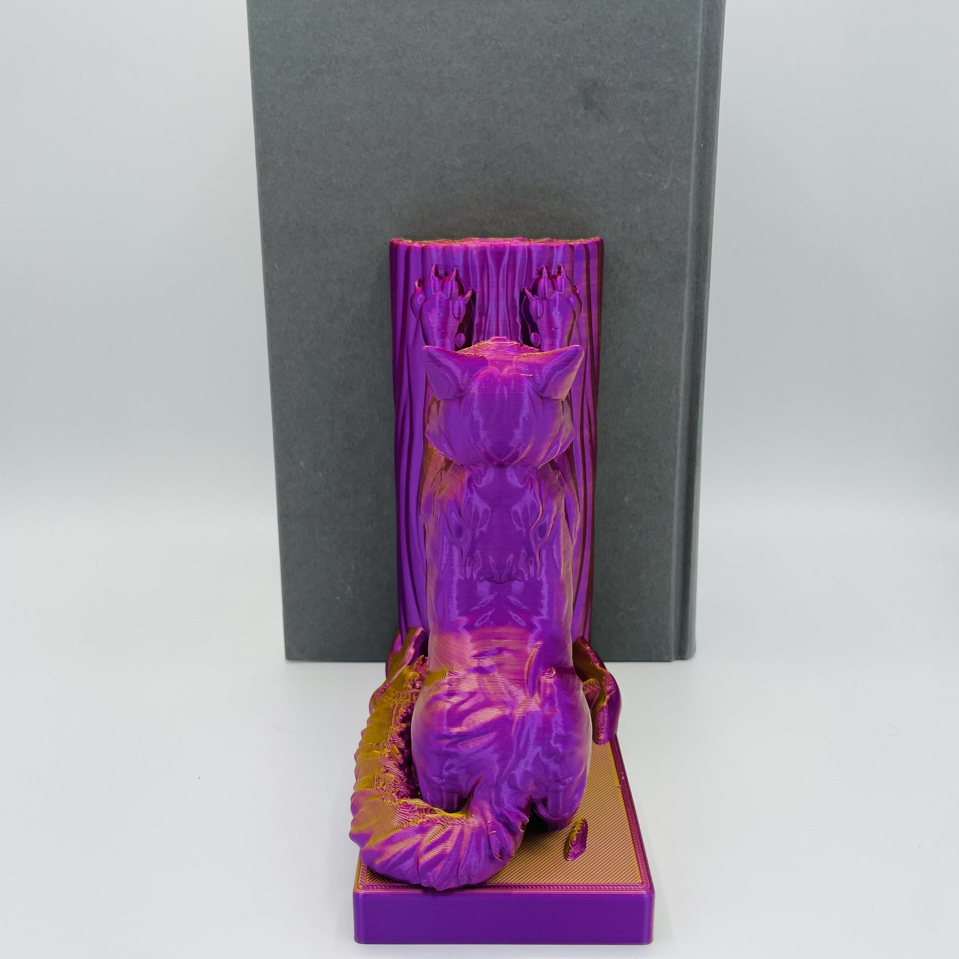 Ansicht des Rückens einer 3D-gedruckten Buchstütze einer süßen, sitzenden Katze in Seitenansicht, die sich mit ihren Vorderpfoten an einen Baum streckt im Farbton Lila-Gold, die Buchstützte steht rechts vom Buch
