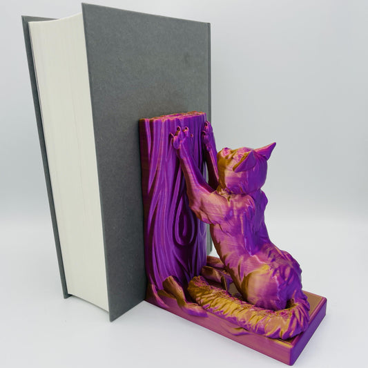 Diagonale Ansicht einer 3D-gedruckten Buchstütze einer süßen, sitzenden Katze in Seitenansicht, die sich mit ihren Vorderpfoten an einen Baum streckt im Farbton Lila-Gold, die Buchstützte steht rechts vom Buch