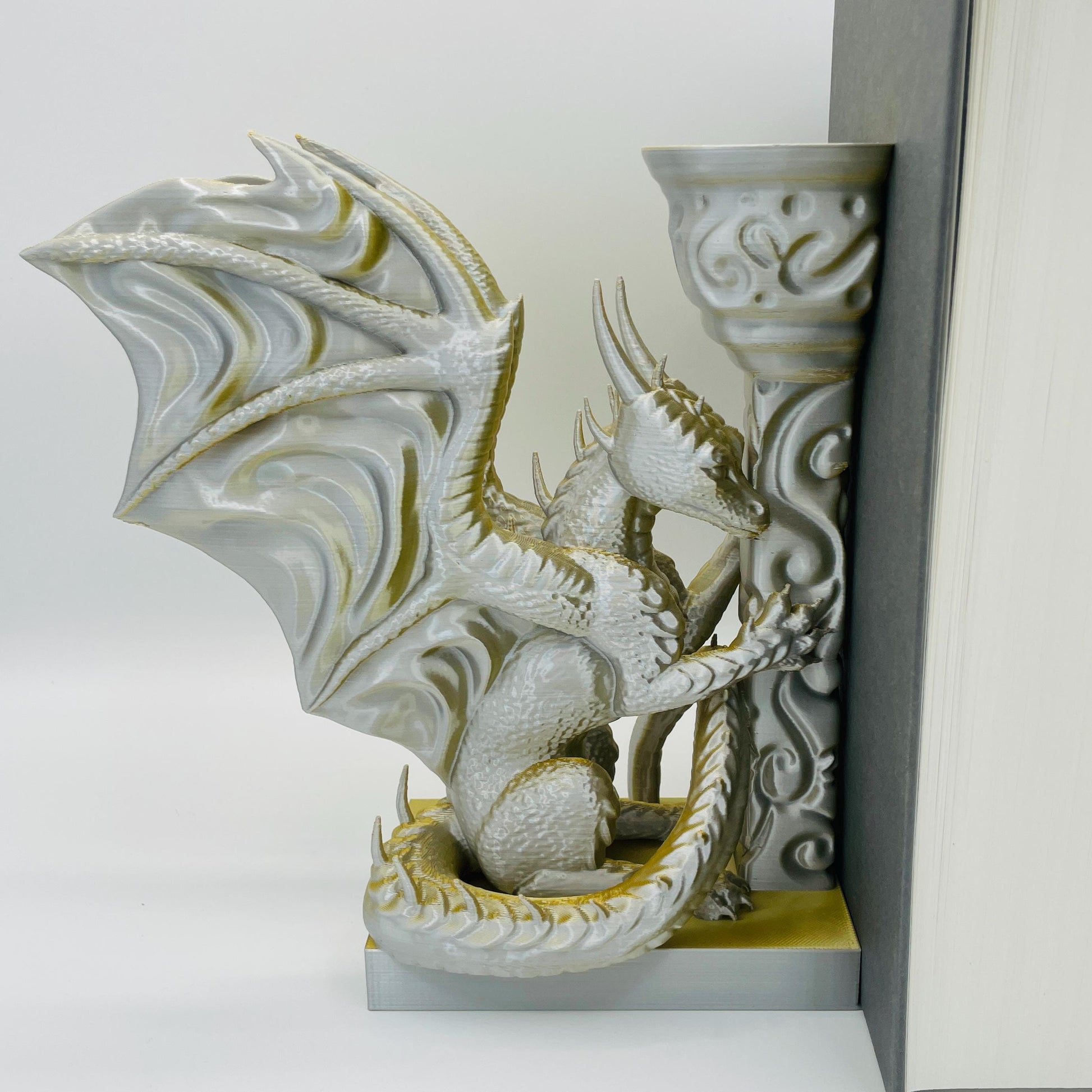 Detailliertere Ansicht einer 3D-gedruckten Buchstütze eines Drachens in Seitenansicht, angelehnt an eine Säule im Farbton Gold-Silber, die Buchstützte steht links vom Buch