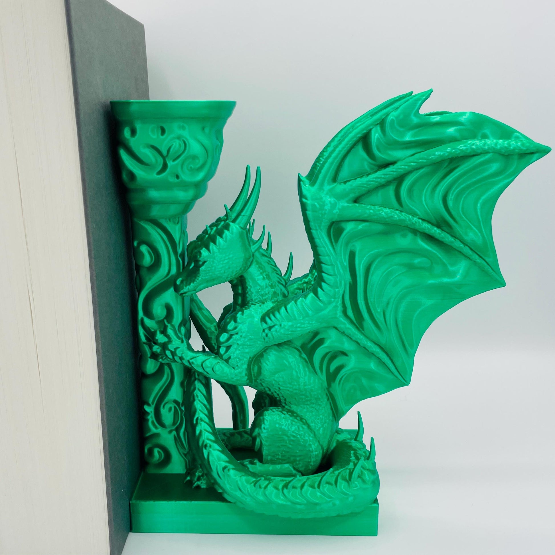 Detailliertere Ansicht einer 3D-gedruckten Buchstütze eines Drachens in Seitenansicht, angelehnt an eine Säule im Farbton Smaragd Grün, die Buchstützte steht rechts vom Buch