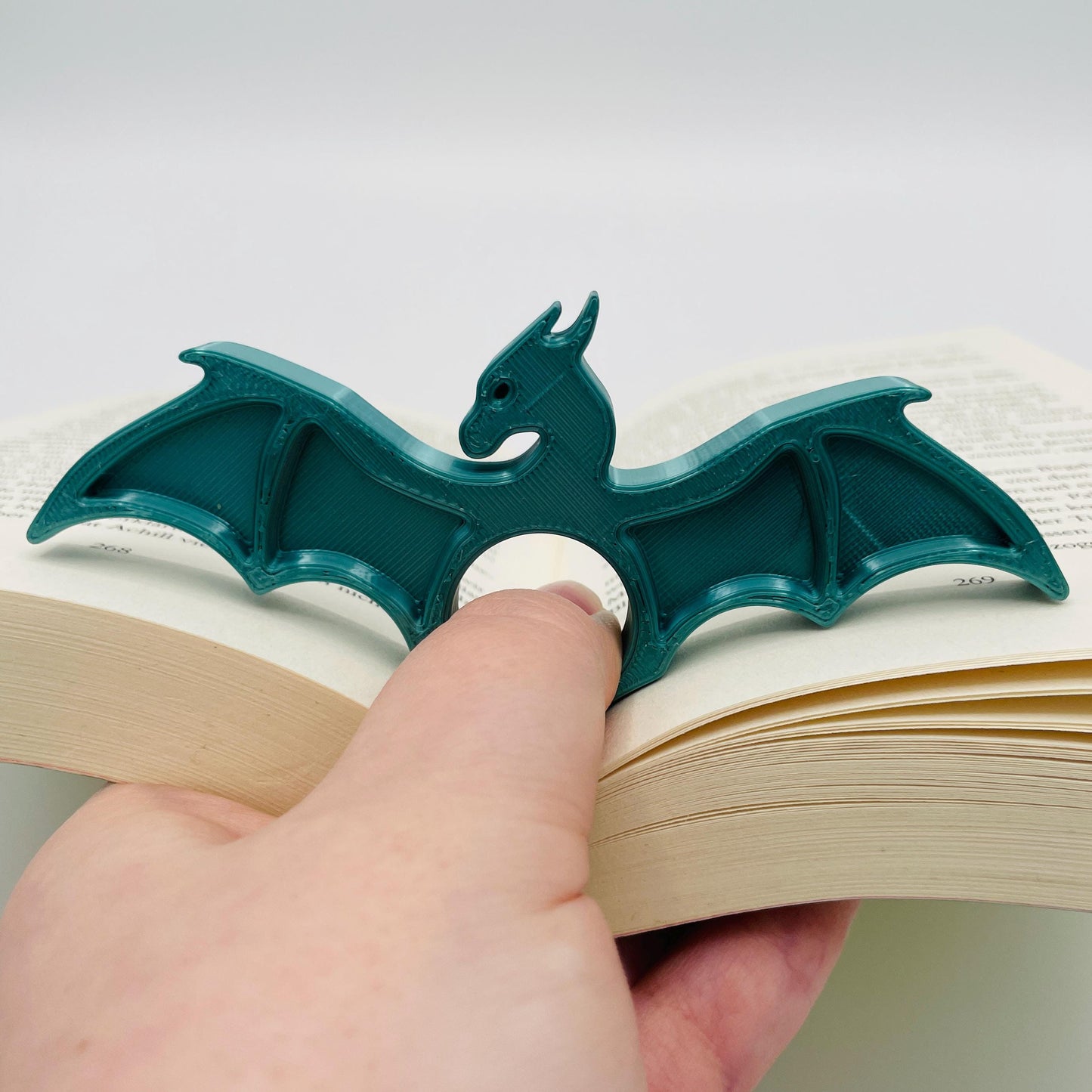 Detailansicht eines 3D-gedruckten Buchseitenhalters in Form eines Drachen mit Lochaussparung für Finger im Farbton Metallic Türkis zur Veranschaulichung in aufgeklapptem Buch