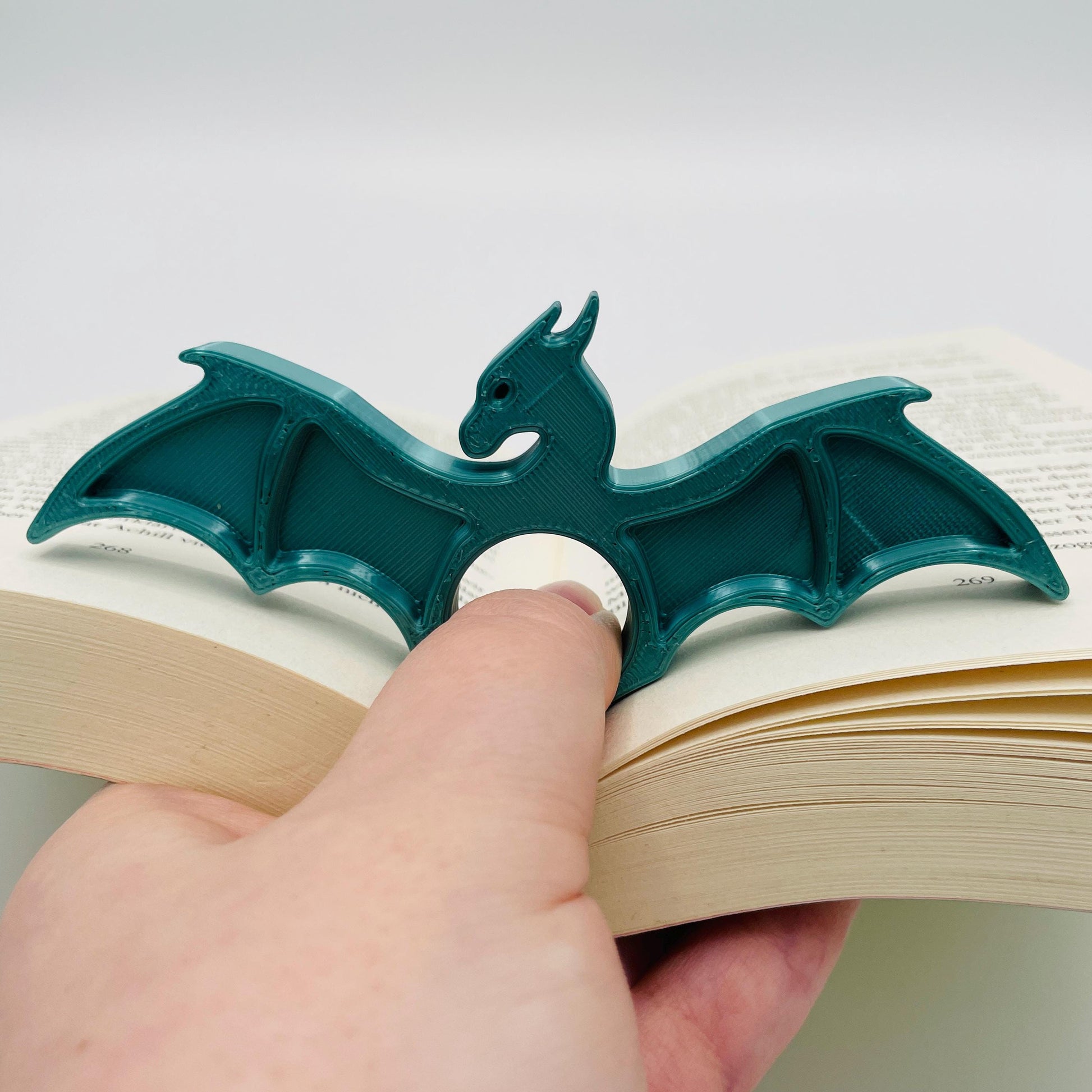 Detailansicht eines 3D-gedruckten Buchseitenhalters in Form eines Drachen mit Lochaussparung für Finger im Farbton Metallic Türkis zur Veranschaulichung in aufgeklapptem Buch