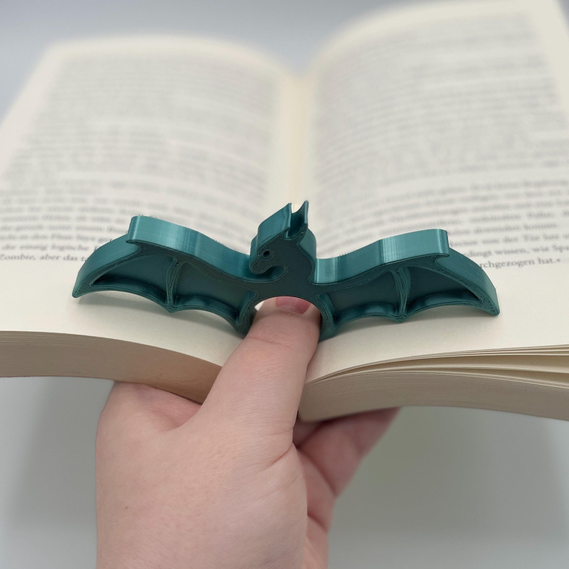 Diagonale Ansicht eines 3D-gedruckten Buchseitenhalters in Form eines Drachen mit Lochaussparung für Finger im Farbton Metallic Türkis zur Veranschaulichung in aufgeklapptem Buch