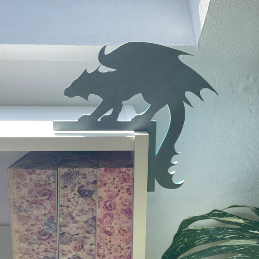 Dekorativer 3D-gedruckter wachende Drache auf einem rechtwinkligen Balken im Farbton Metallic Olivgrün mit texturierter Oberfläche der auf der Ecke eines Bücherregals sitzt.