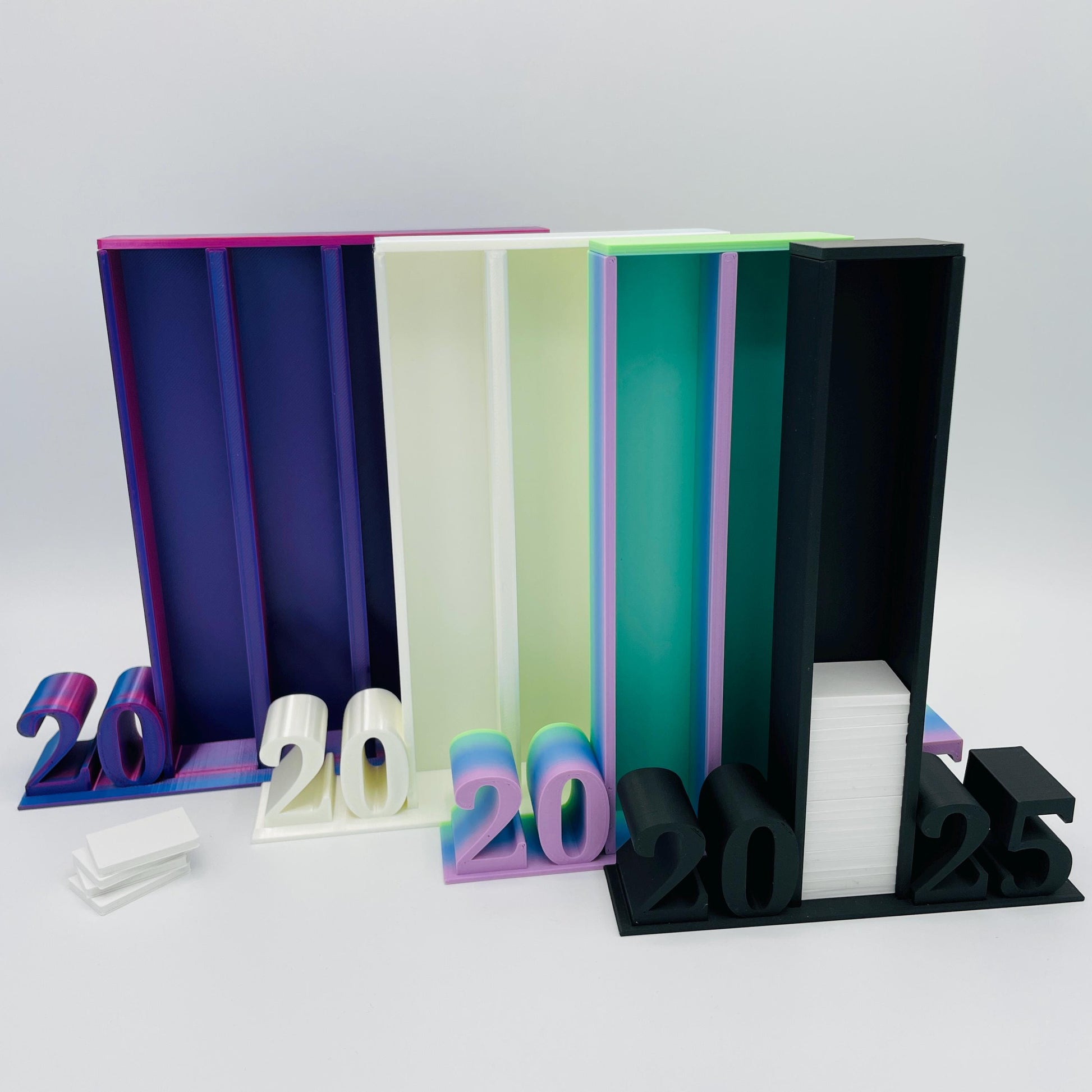 Frontansicht 3D-gedruckte Lesetürme mit 2025 Schrift im Vergleich, 4 Spalten in Blau-Pink-Lila, 3 Spalten in Perlmutt Weiß, 2 Spalten in RB Matt Regenbogen-Farbverlauf und 1 Spalte in Matt Schwarz für je 50 Mini-Bücher