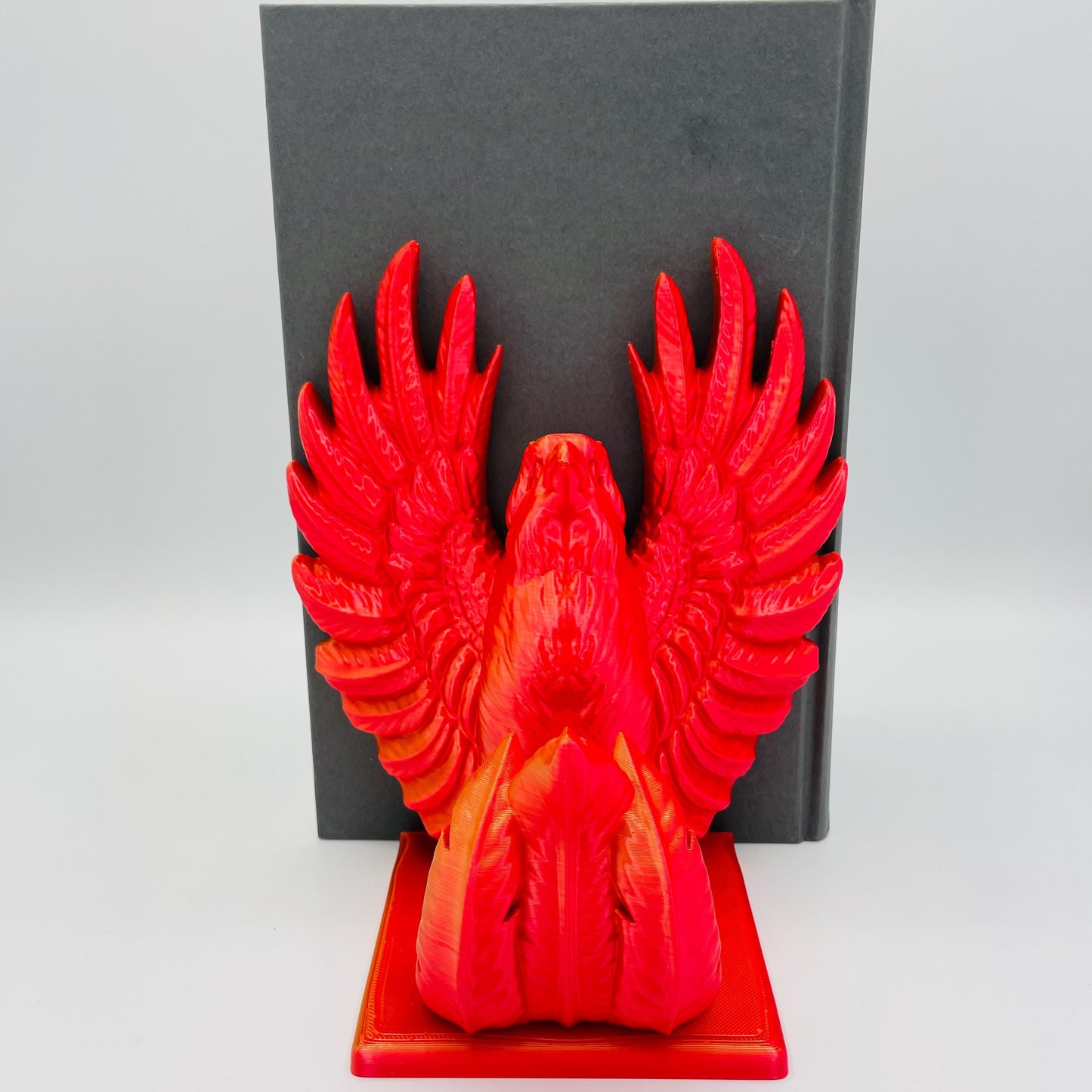 Frontale Ansicht einer 3D-gedruckten Buchstütze eines stolzen Phönix mit gestreckten Flügeln im Farbton Rot-Gold, die Buchstützte steht rechts vom Buch