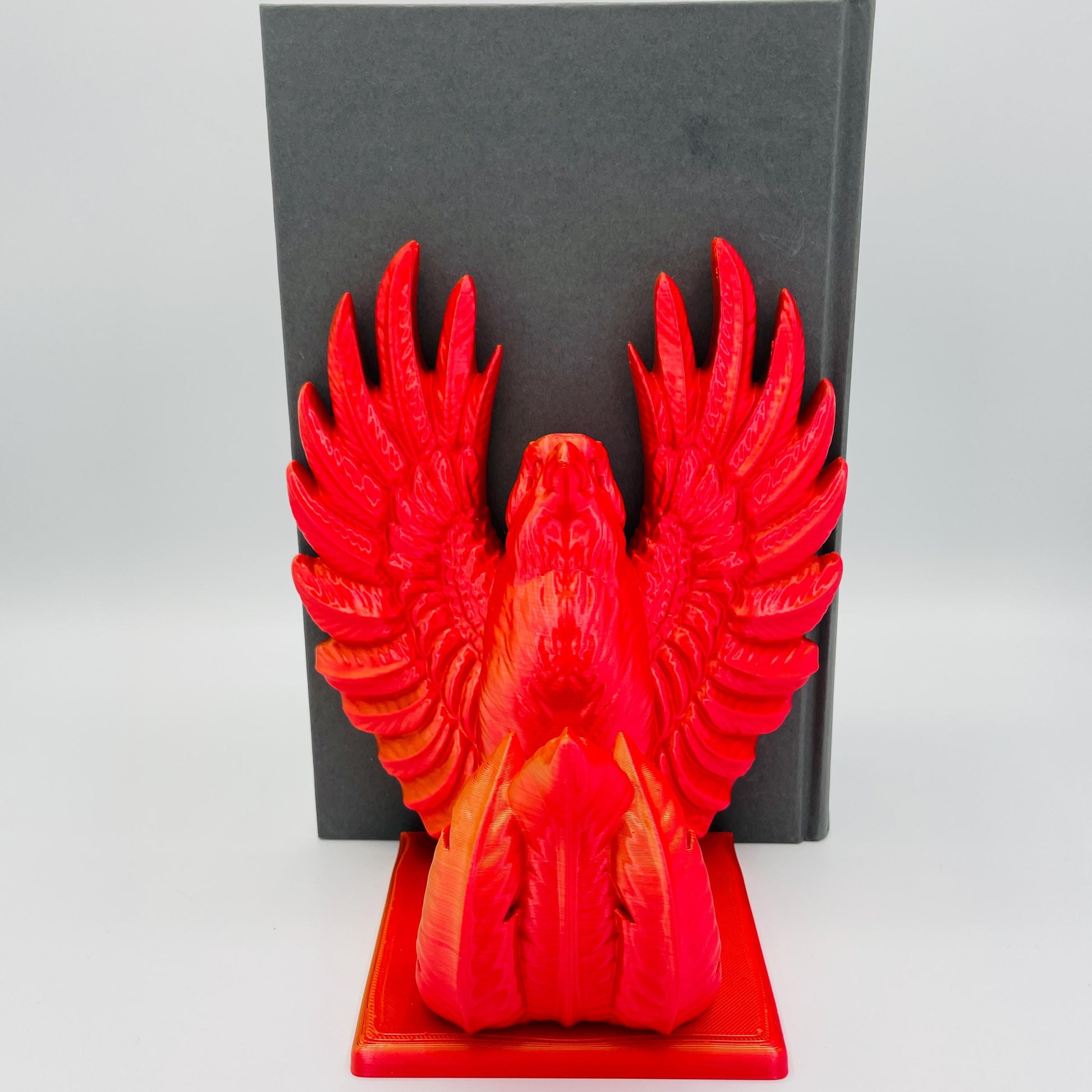 Frontale Ansicht einer 3D-gedruckten Buchstütze eines stolzen Phönix mit gestreckten Flügeln im Farbton Rot-Gold, die Buchstützte steht rechts vom Buch