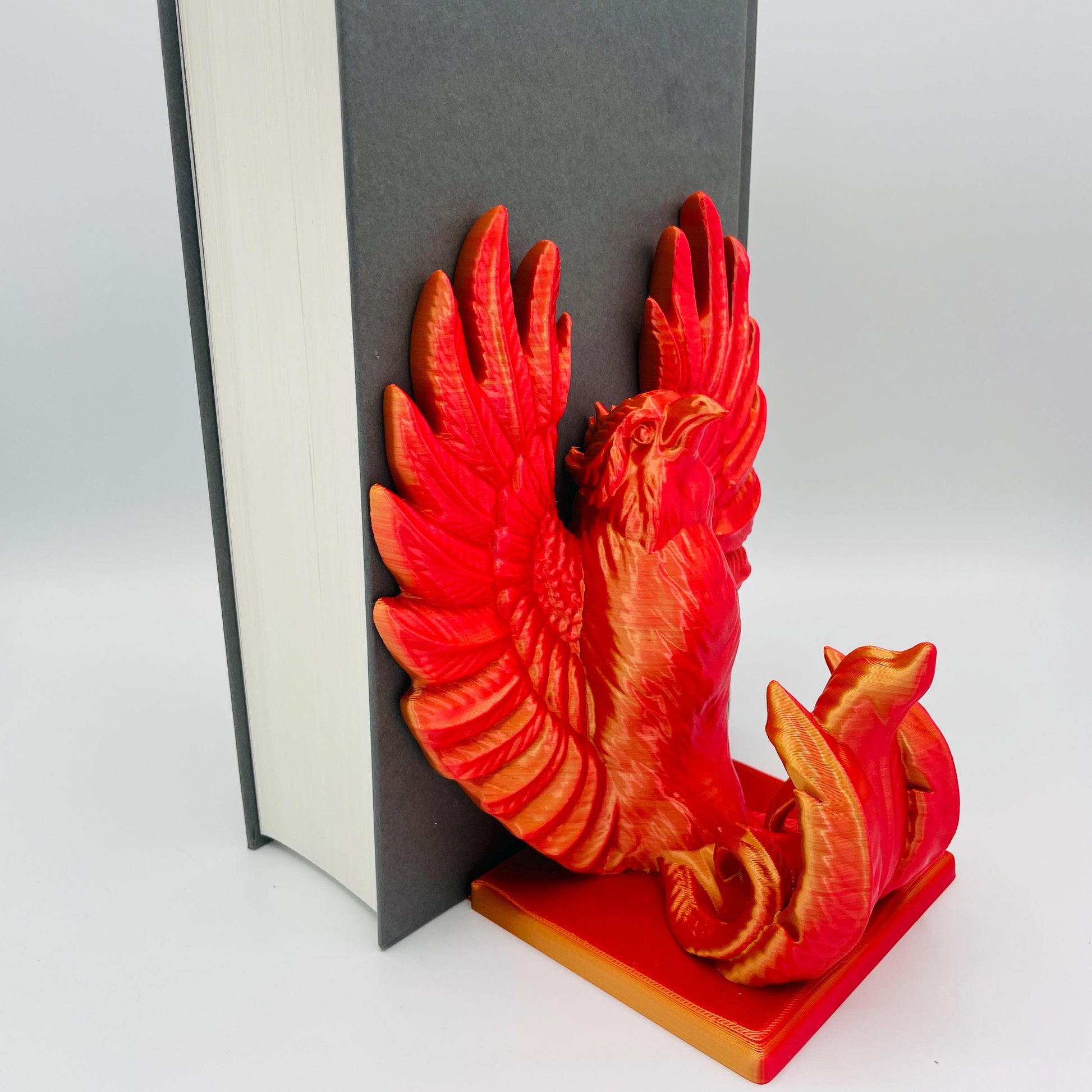 Diagonale Ansicht einer 3D-gedruckten Buchstütze eines stolzen Phönix mit gestreckten Flügeln im Farbton Rot-Gold, die Buchstützte steht rechts vom Buch