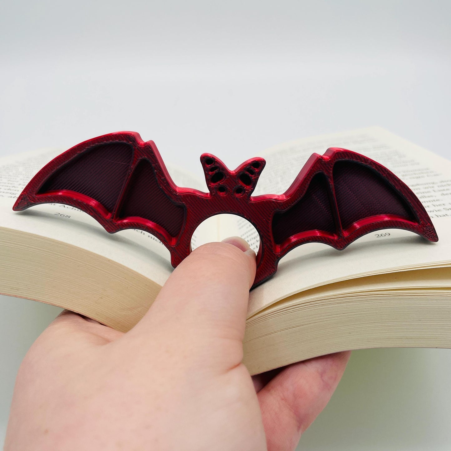Diagonale Ansicht eines 3D-gedruckten Buchseitenhalters in Form einer Fledermaus mit Lochaussparung für Finger im Farbton Rot-Schwarz zur Veranschaulichung in aufgeklapptem Buch