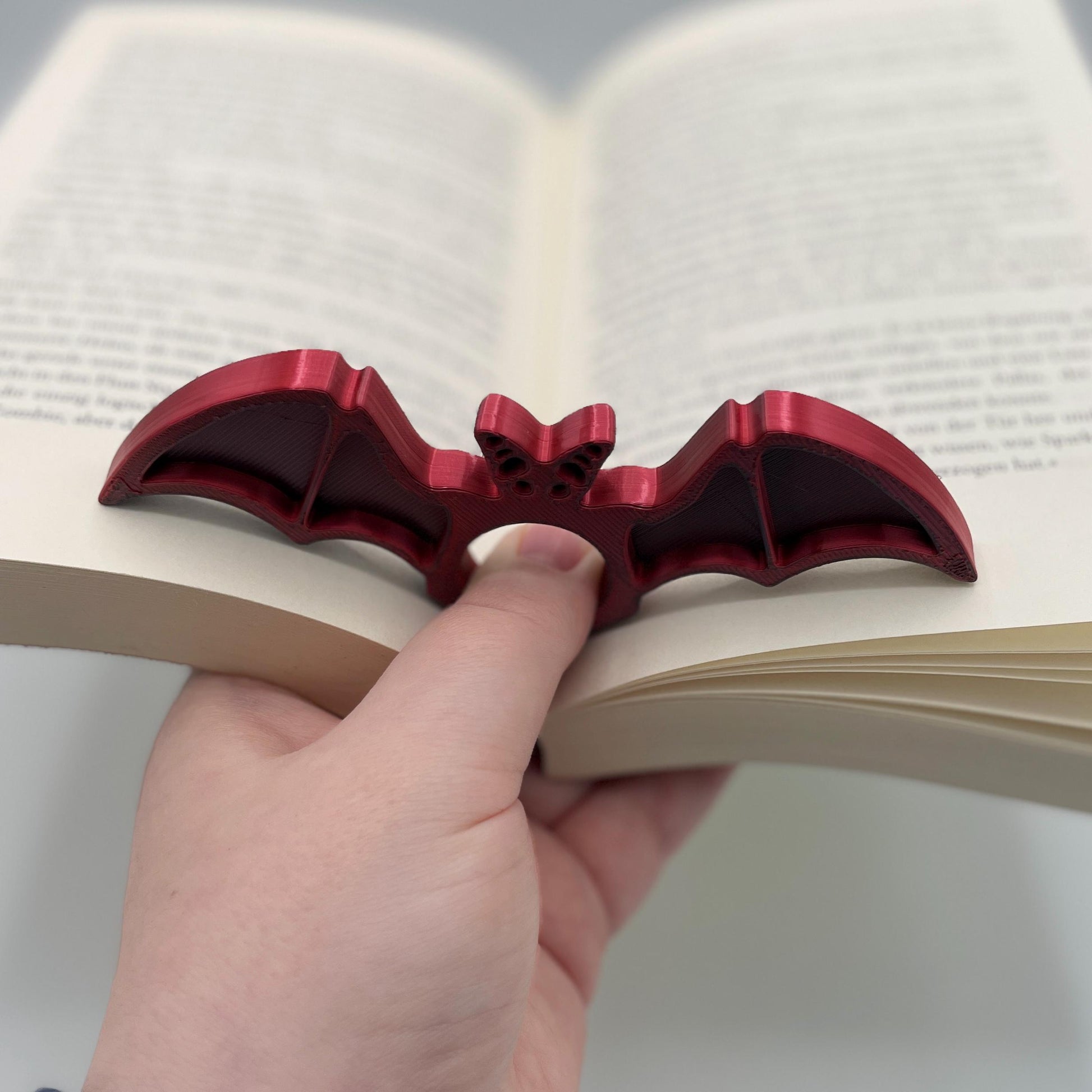Diagonale Ansicht eines 3D-gedruckten Buchseitenhalters in Form einer Fledermaus mit Lochaussparung für Finger im Farbton Rot-Schwarz zur Veranschaulichung in aufgeklapptem Buch