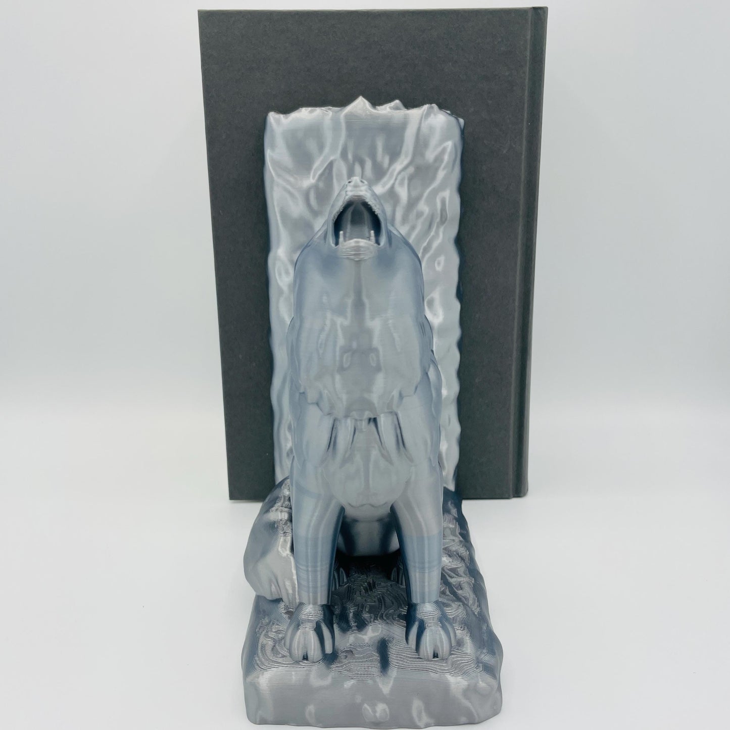 Frontale Ansicht einer 3D-gedruckten Buchstütze eines starken, sitzenden Wolfes, der zum Himmel heult/jault, im Farbton Silber, die Buchstützte steht rechts vom Buch