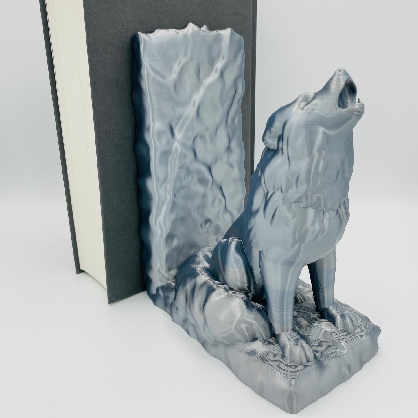 Diagonale Ansicht einer 3D-gedruckten Buchstütze eines starken, sitzenden Wolfes, der zum Himmel heult/jault, im Farbton Silber, die Buchstützte steht rechts vom Buch